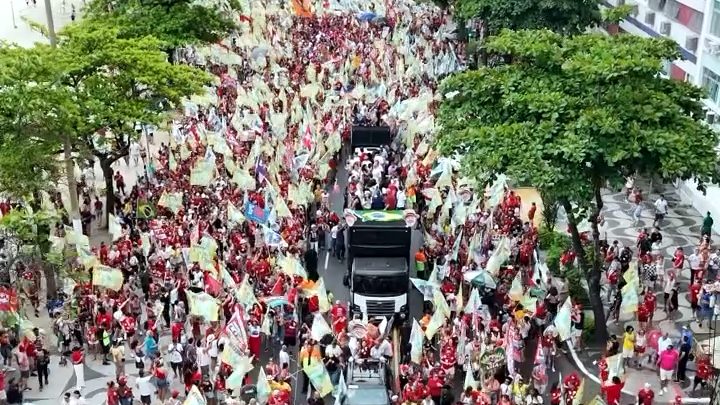 Vista do alto de uma avenida arborizada tomada por manifestantes, a maioria de camisa vermelha, com bandeiras claras e um caminhão de som no centro.