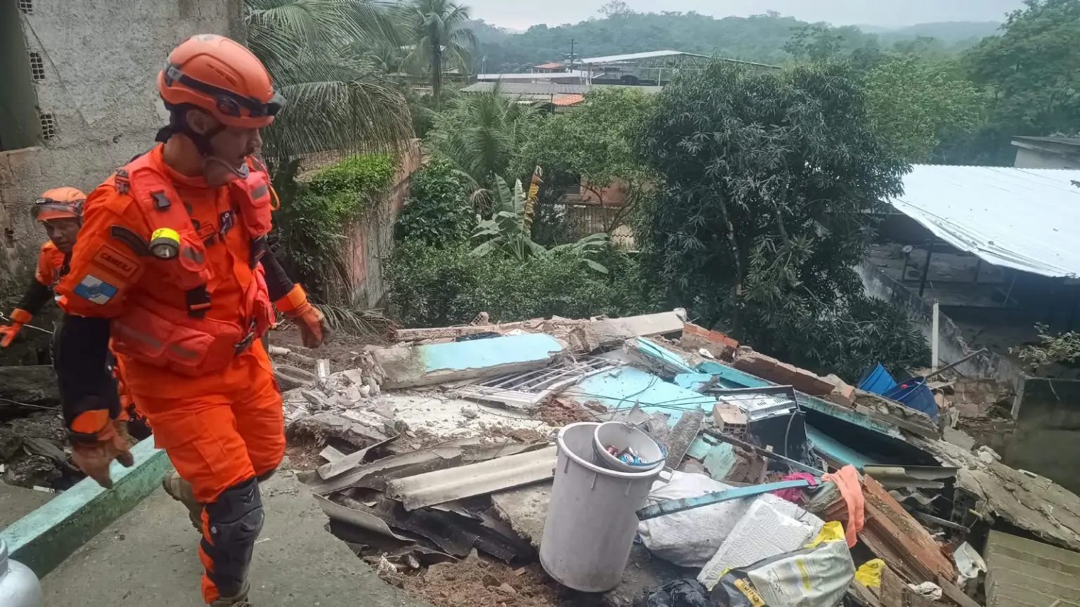 Duas pessoas de uniforme e capacete laranja caminham por um piso de concreto ao lado de escombros, com pedaços de parede azul-clara, tijolos, telhas, um balde branco e sacos. Ao fundo, árvores e morros cobertos de vegetação.