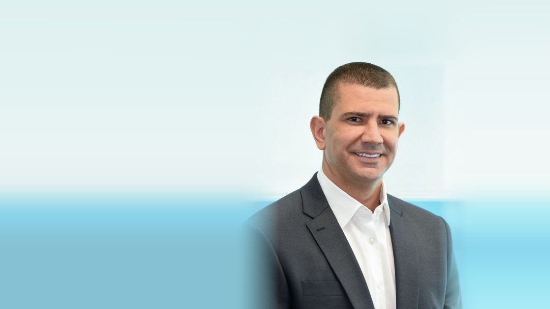 Douglas Ruas sorri em retrato, de terno cinza-escuro e camisa branca, diante de um fundo claro com uma faixa azul.