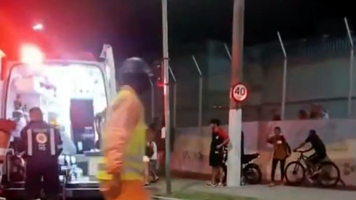 Ambulância do Samu com as portas abertas e as luzes acesas numa avenida à noite, com socorristas e pessoas junto a um muro com grade.