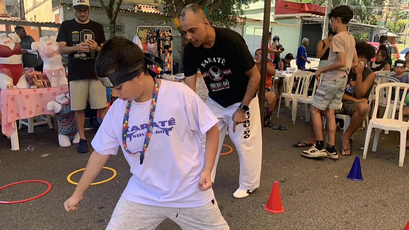 Menino de camiseta branca do karatê e faixa na cabeça, em posição de luta numa rua; ao lado, um professor de calça branca o acompanha, e o público assiste ao fundo.