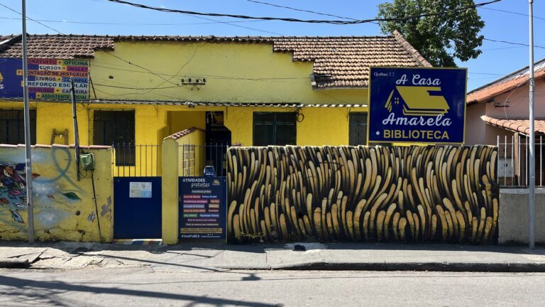 Casa amarela de um andar com a placa azul da Biblioteca A Casa Amarela, uma faixa com a lista de atividades gratuitas e um muro grafitado em amarelo e preto.