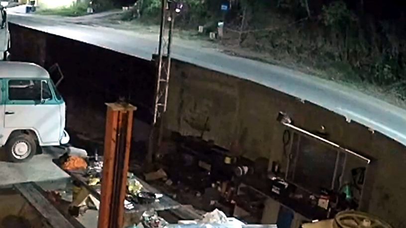 Trecho de estrada em curva registrado à noite por câmera de segurança, com vegetação ao redor e pouca iluminação.