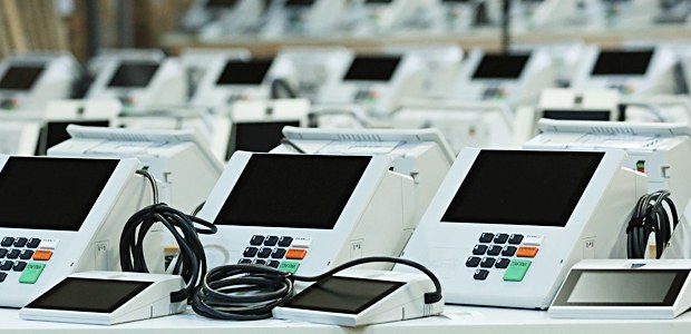 Dezenas de urnas eletrônicas brancas enfileiradas sobre mesas, com os cabos enrolados ao lado de cada uma