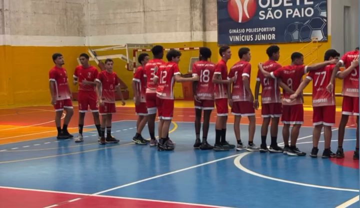 Jogadores de camisa vermelha perfilados lado a lado numa quadra azul, com uma faixa do colégio ao fundo.
