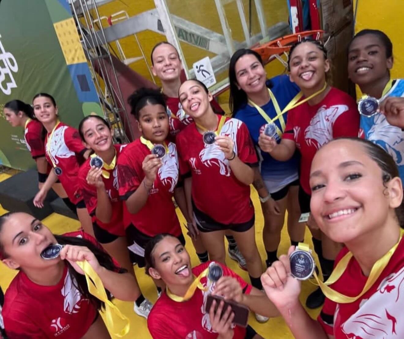 Jogadoras de camisa vermelha sorriem para a câmera e mostram medalhas com fitas amarelas.