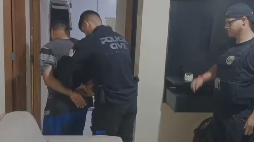 Policial civil conduz homem algemado, de costas, pelo corredor de uma casa; outro agente observa ao lado.