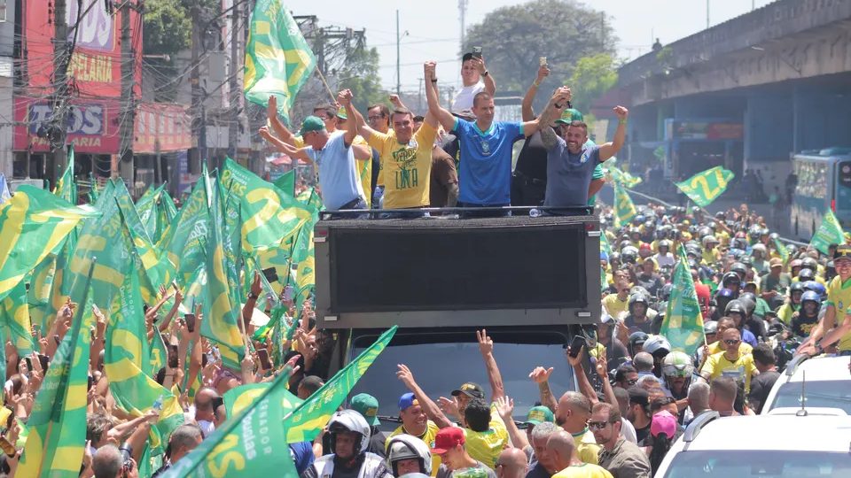 Homens acenam de cima de um caminhão no meio de uma multidão de camisas verdes e amarelas, com bandeiras verdes com o número 22 e motociclistas ao redor.