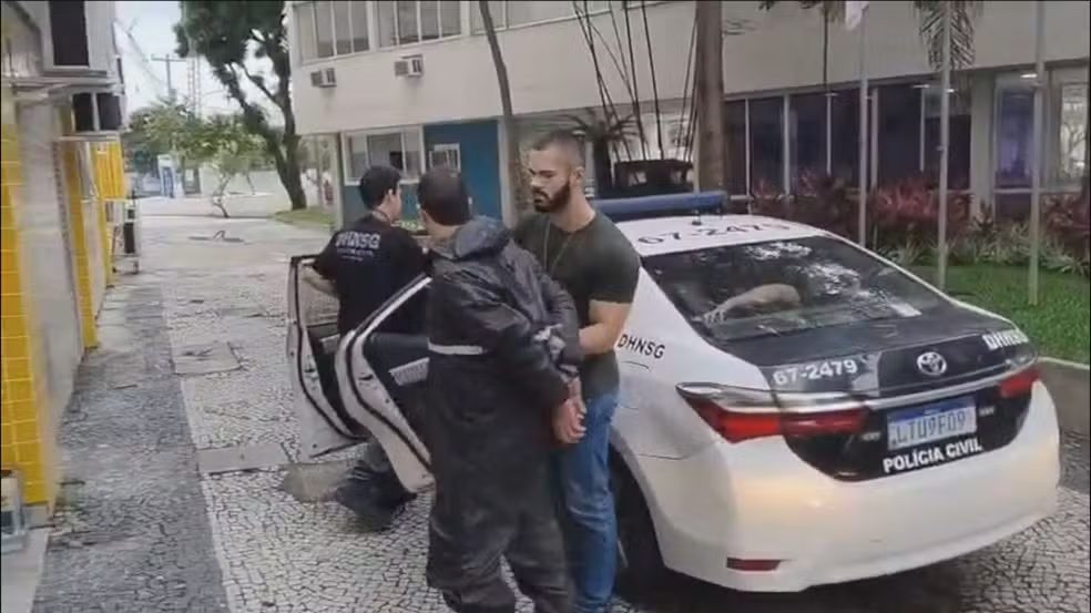 Homem algemado é conduzido por agentes da Polícia Civil a viatura da DHNSG em área externa de prédio.