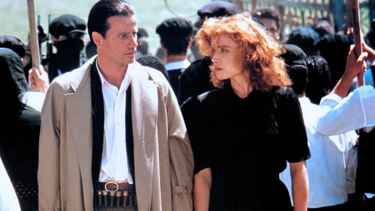 Cena de filme: um homem de sobretudo bege e cinto de munição caminha ao lado de uma mulher ruiva de vestido preto, no meio de uma multidão com bastões erguidos.