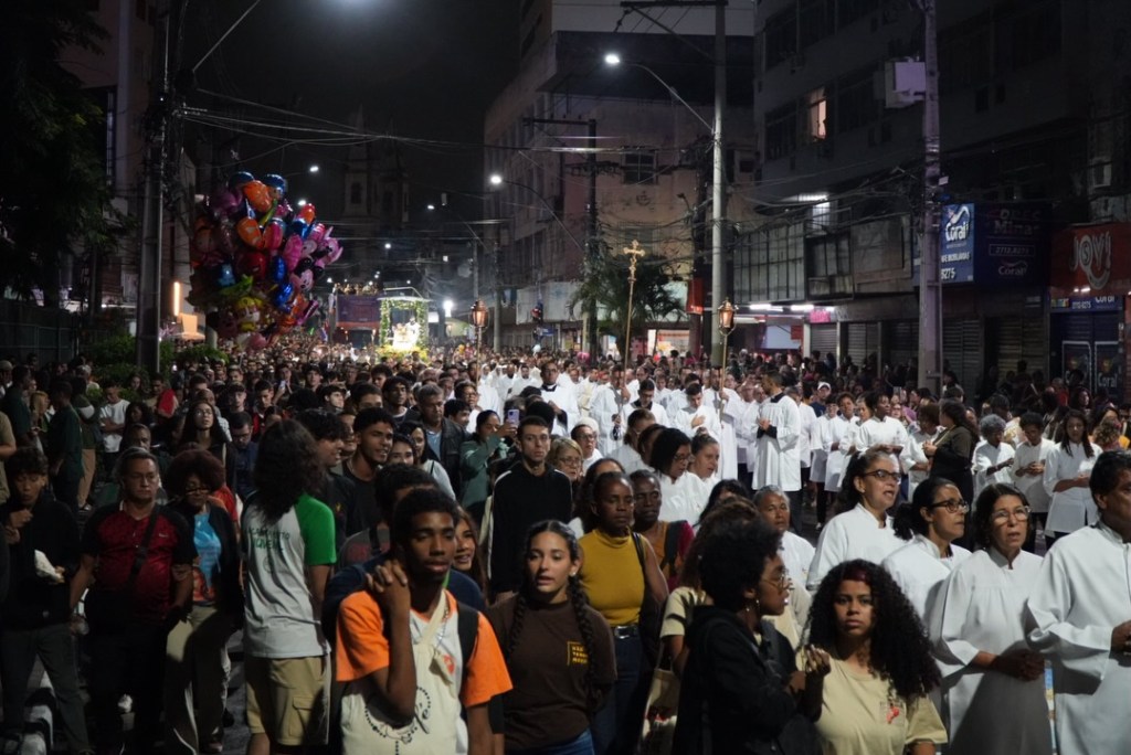 Uma grande multidão participa de um evento noturno, com pessoas vestindo roupas brancas e segurando balões coloridos enquanto caminham pela rua.