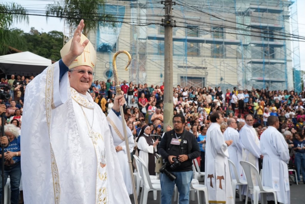 Um bispo acenando para a multidão em um evento religioso, cercado por padres, com um grande público ao fundo.
