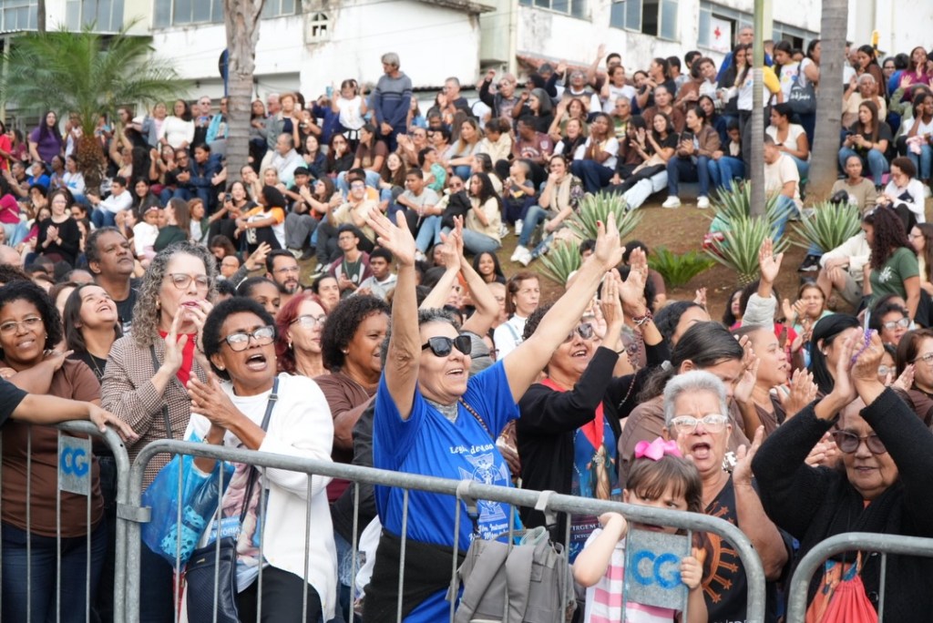 Um grande grupo de pessoas aplaudindo e vibrando em um evento ao ar livre, com pessoas em várias faixas etárias. Algumas pessoas estão de pé, outras estão sentadas, e todas mostram expressões de alegria e entusiasmo.