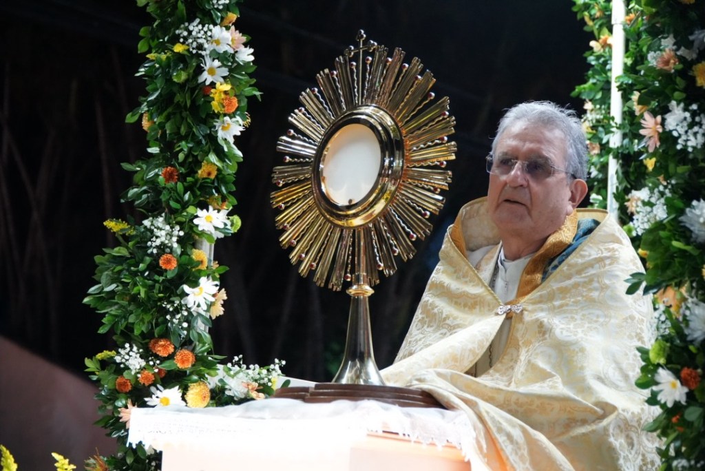Um religioso sentado em um altar decorado com flores, apresentando um ostensório dourado com a hóstia sagrada.